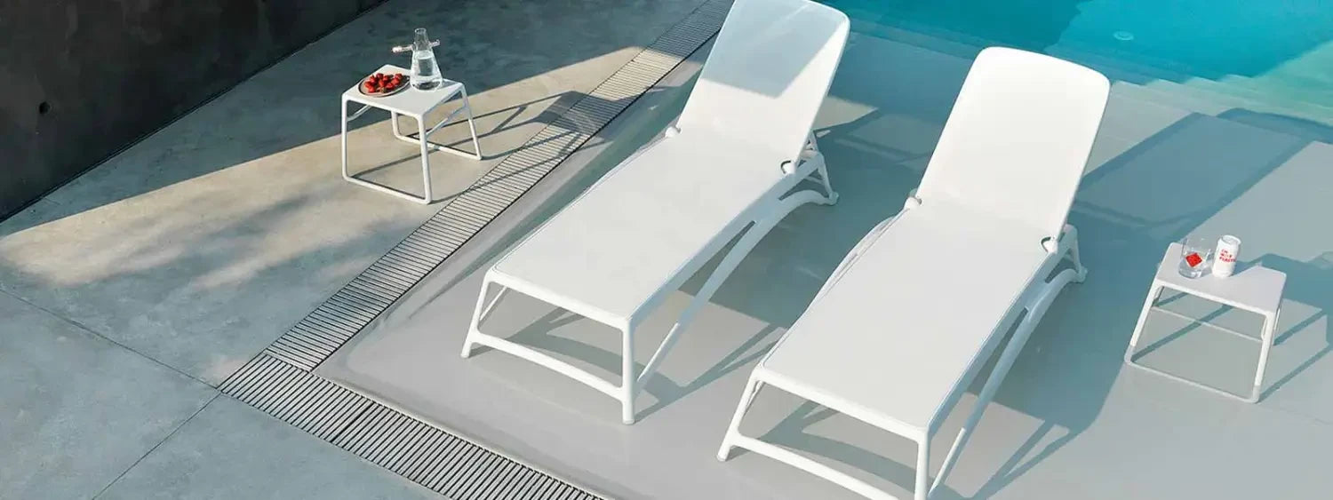 White outdoor sunloungers with matching side tables by the poolside.